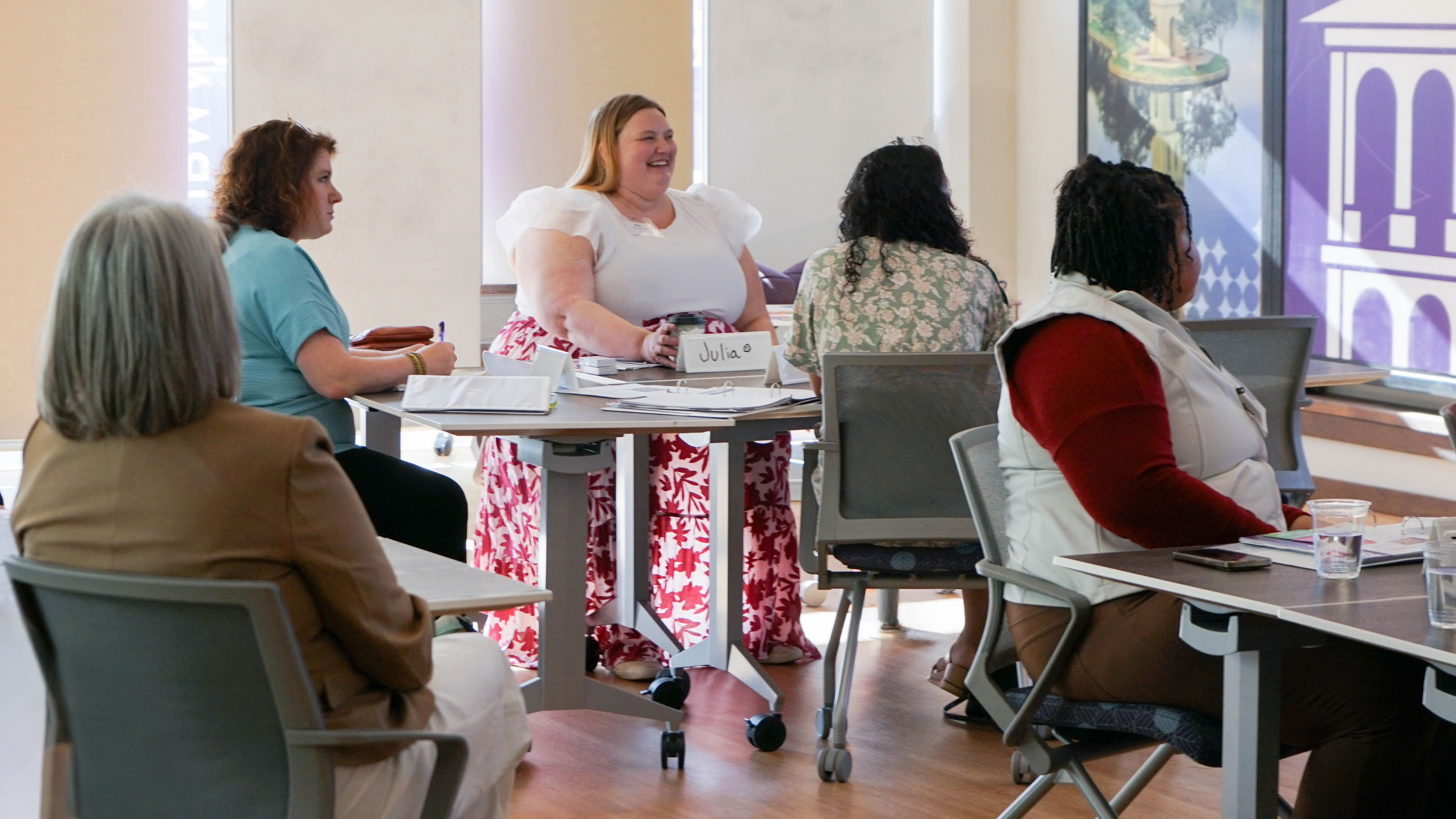 A group of adults sit around small tables in a bright classroom, with notebooks, papers, and cups on the tables; one person at the center sits at a round table with a visible name card reading “Julia” while others face forward or toward each other.