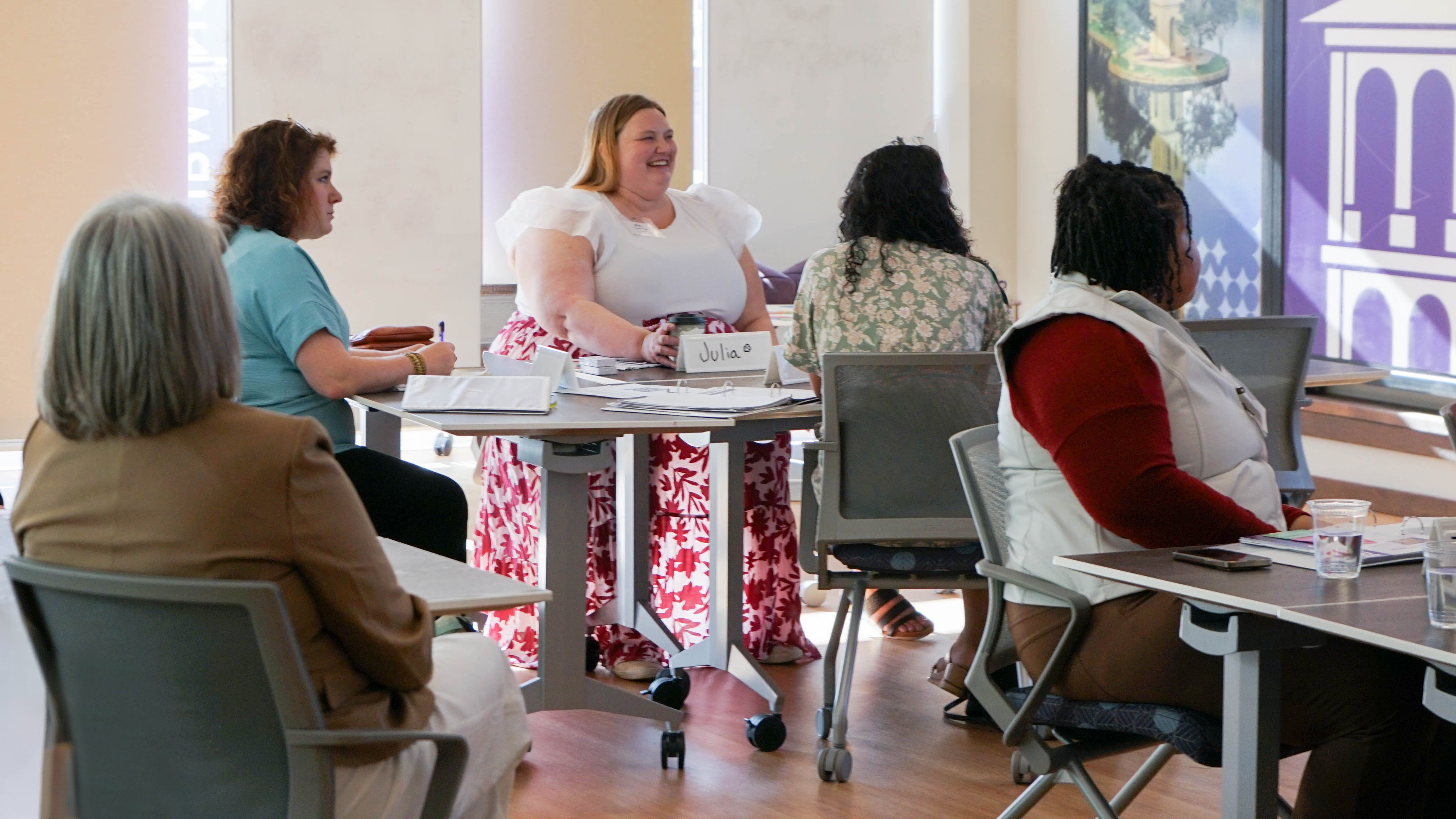A group of adults sit around small tables in a bright classroom, with notebooks, papers, and cups on the tables; one person at the center sits at a round table with a visible name card reading “Julia” while others face forward or toward each other.